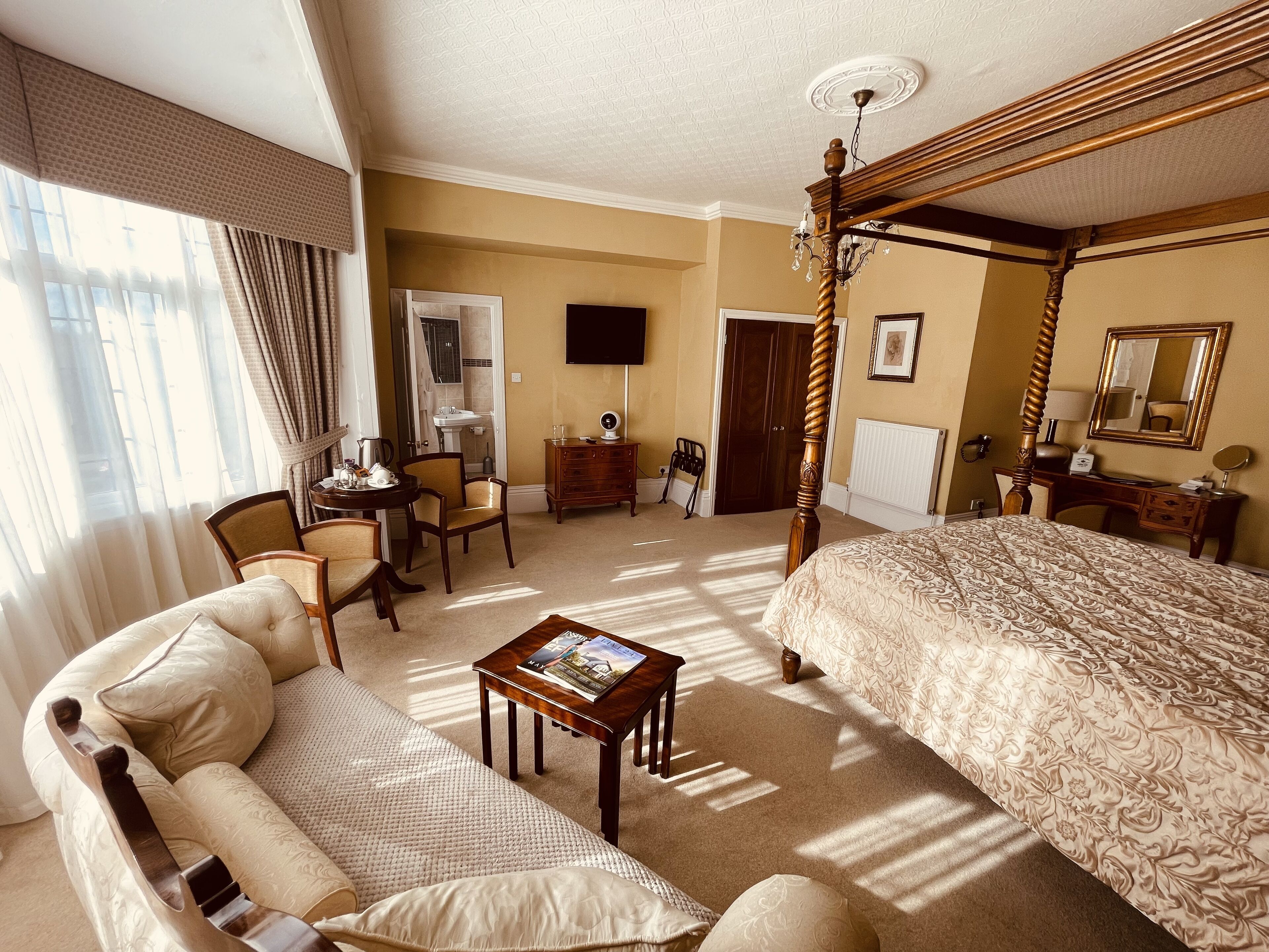 A hotel bedroom with a four-poster bed, a sitting area with a sofa and armchairs, a desk with a chair, and a bathroom with a sink and mirror.