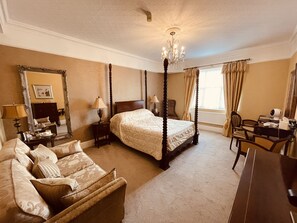 A spacious bedroom with a four-poster bed, a sitting area with a sofa, a desk, and a large mirror.