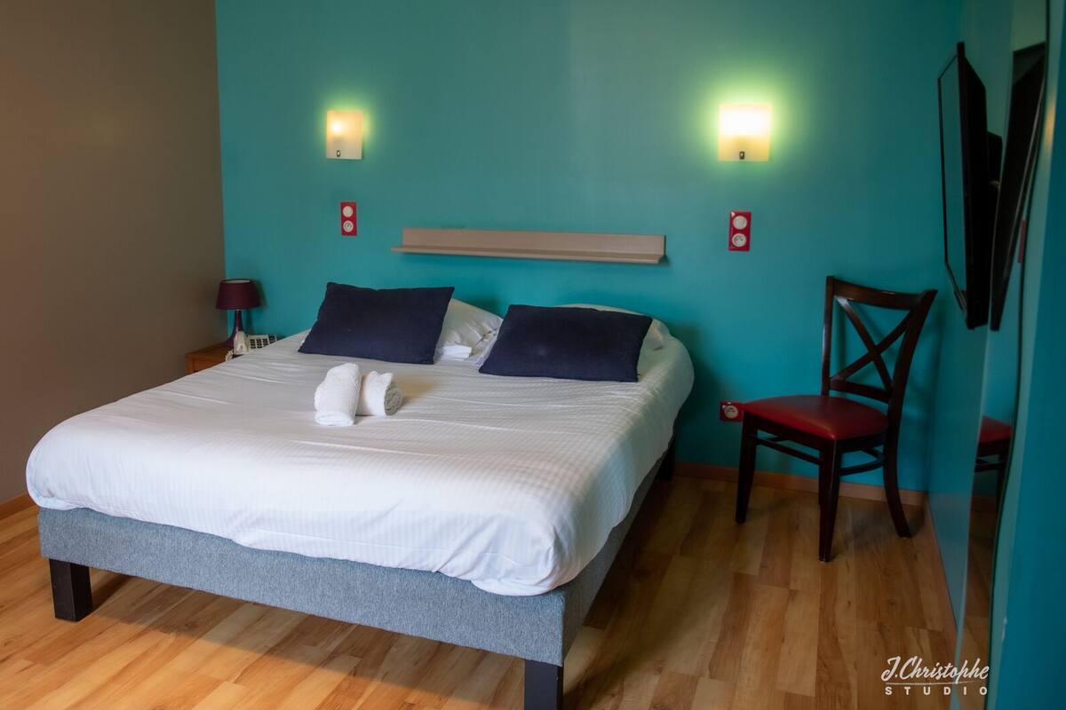A neatly made bed with white linens and two dark pillows, a wooden nightstand with a lamp, a red chair, and a teal-colored wall with two wall-mounted lights.