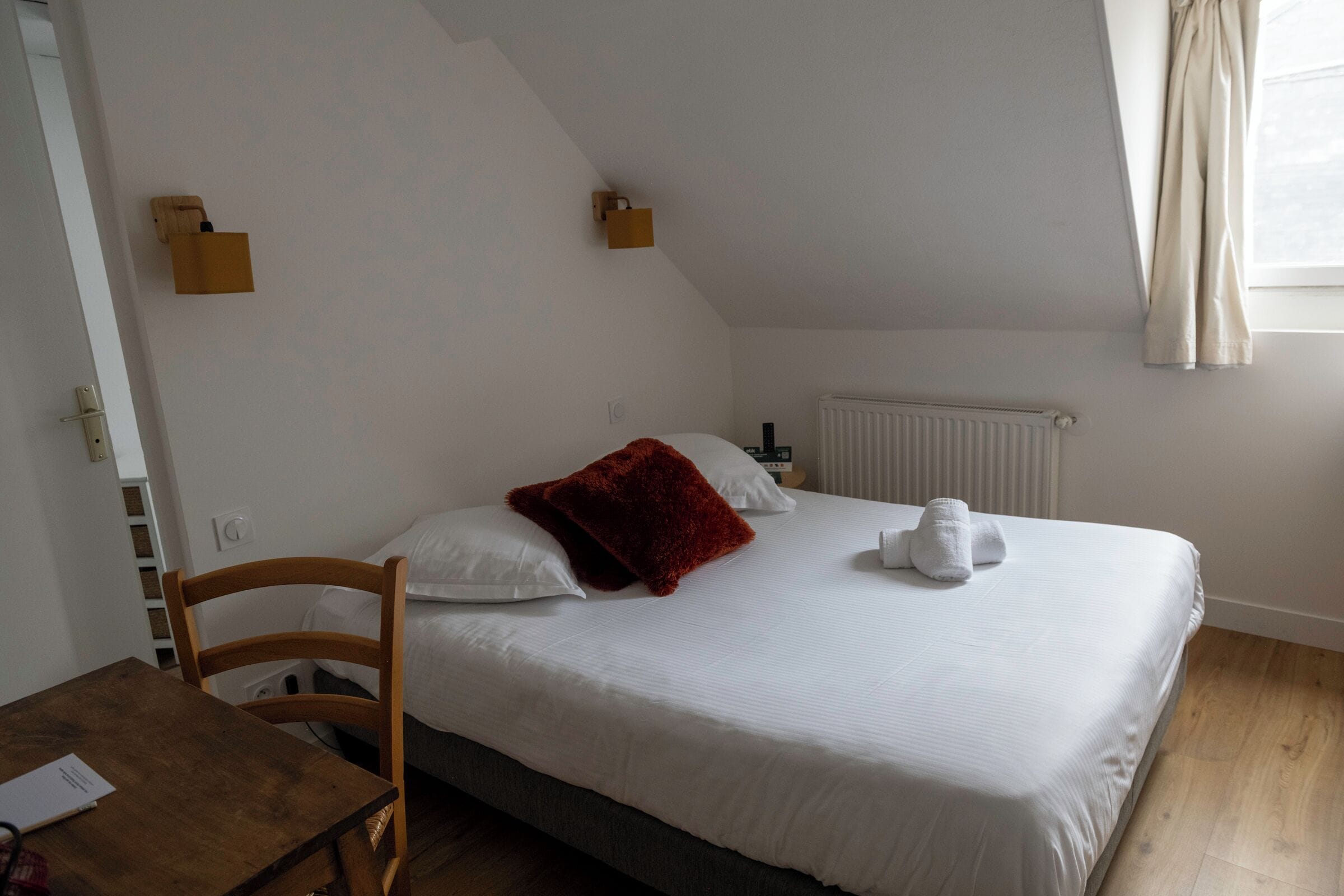 Een eenpersoonsbed met witte lakens en een rode sierkussen in een kamer met een houten vloer en witte muren.