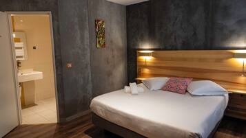 A hotel room with a bed, a nightstand, a wall-mounted lamp, a painting, and a bathroom with a sink and mirror.