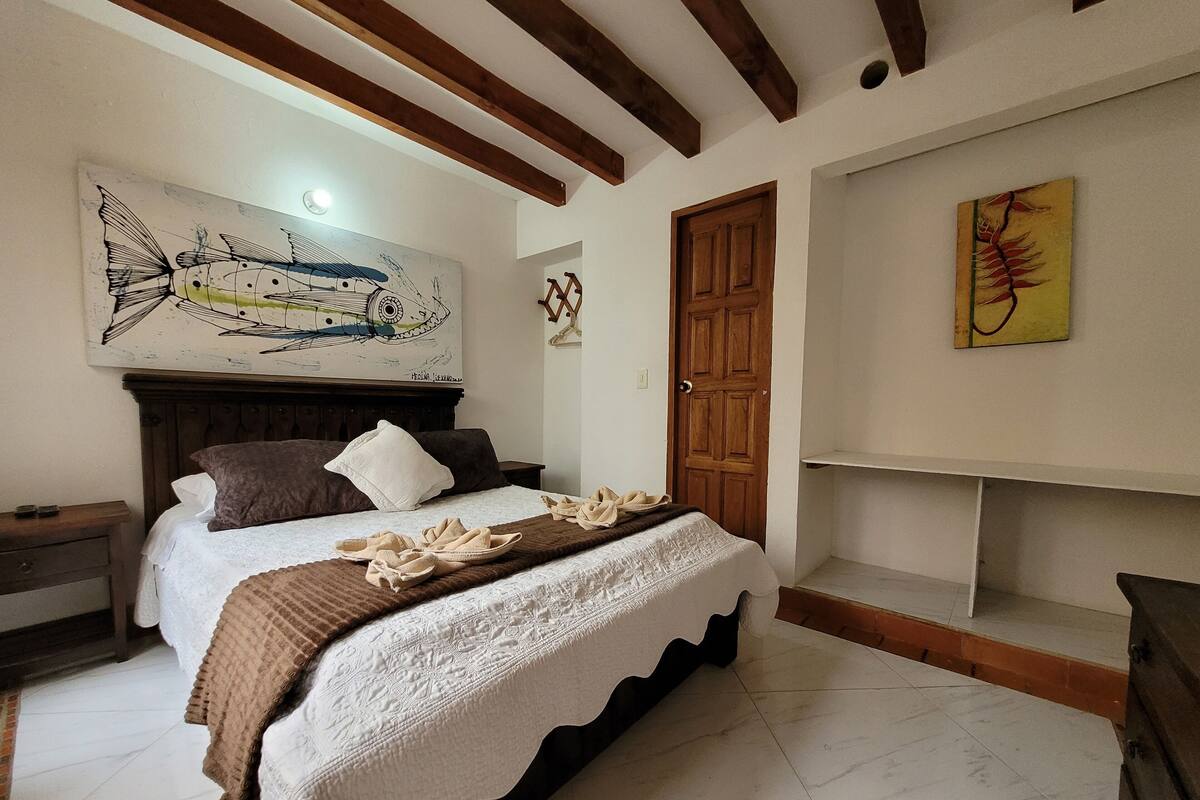 Un dormitorio con cama, cabecera de madera, un cuadro sobre la cama, una puerta de madera y una estantería con un solo objeto.