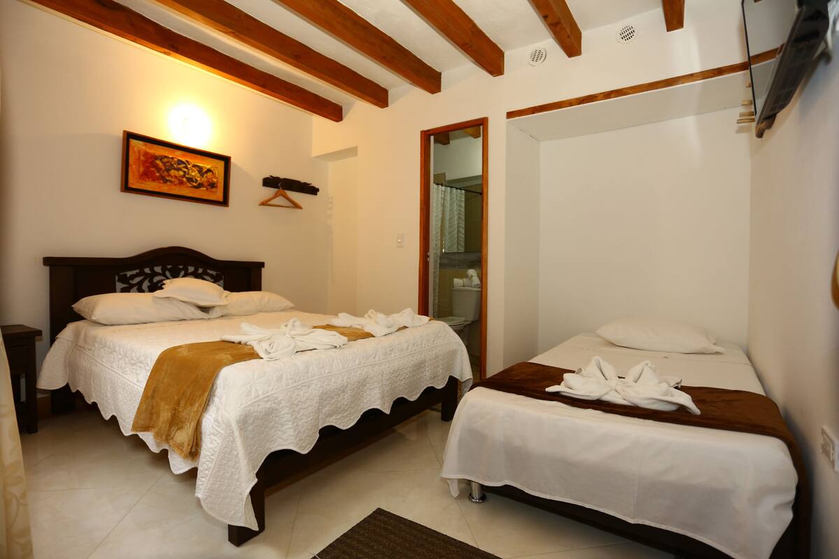 Un dormitorio con dos camas, techo de madera, un cuadro en la pared y un baño visible a través de una puerta abierta.