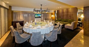 Uma área de jantar com uma mesa redonda posta para seis pessoas, rodeada por seis cadeiras cinzas estofadas. Uma televisão está instalada na parede acima da mesa.