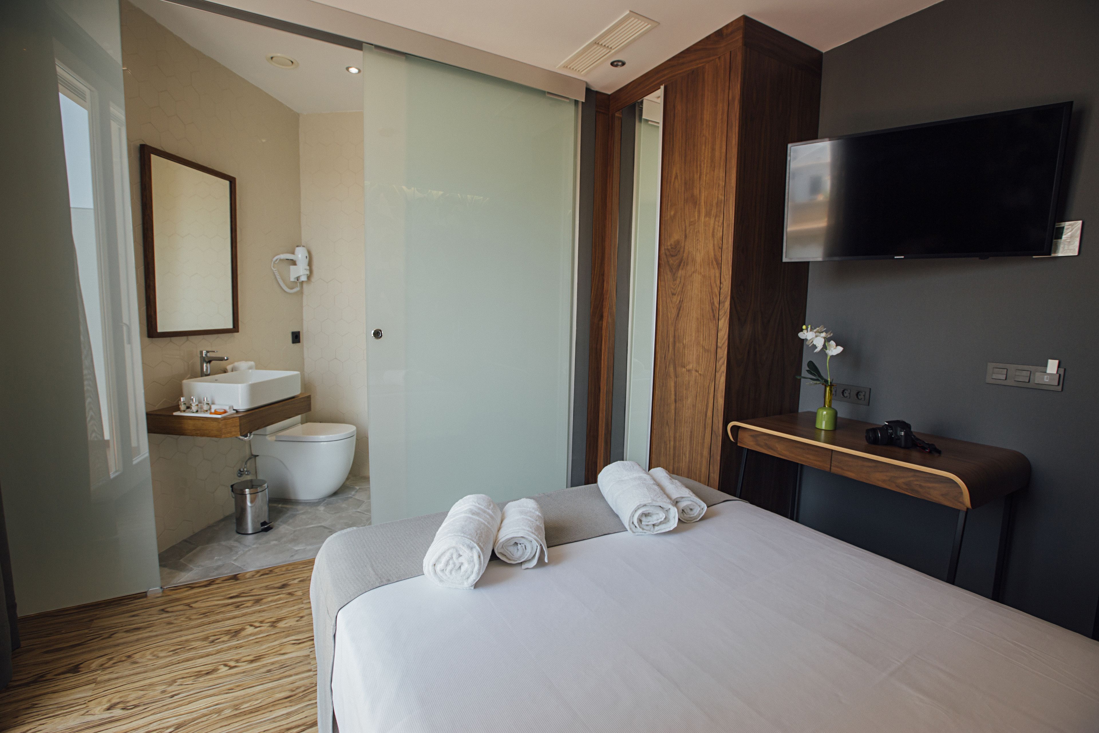 A modern hotel room with a large bed, a wooden desk, a flat-screen TV, a bathroom with a toilet and sink, and a mirror.