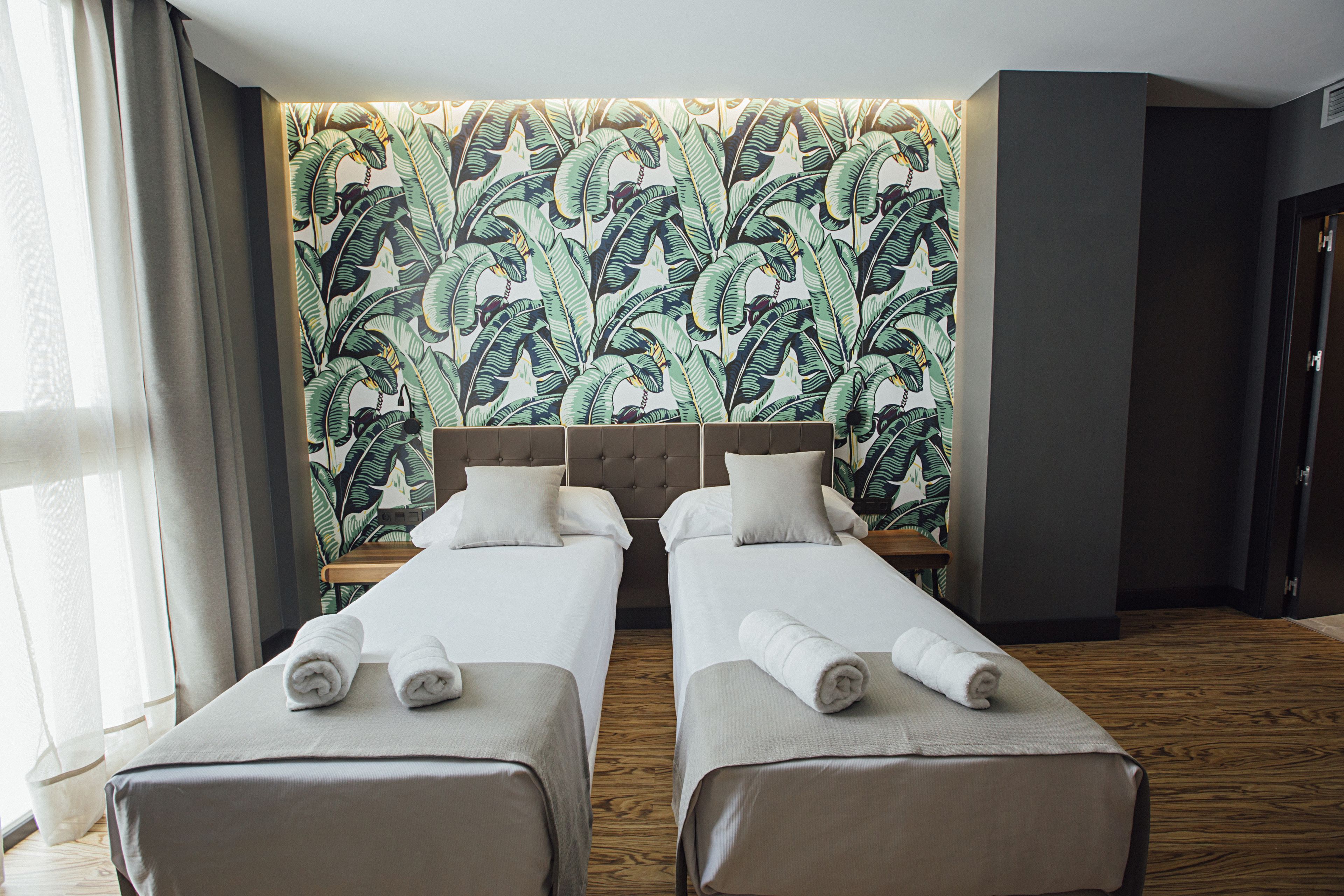 Une chambre d’hôtel avec deux lits simples, une grande fenêtre avec des rideaux légers et un mur orné d’un motif de feuilles tropicales.