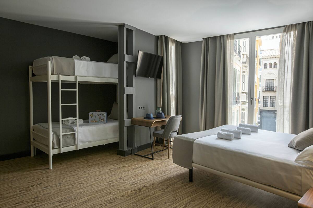 Una habitación de hotel con literas, un escritorio y una silla, un televisor y un ventanal con cortinas.
