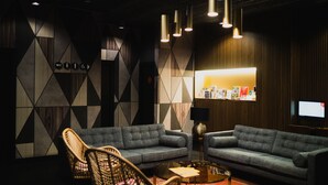 A modern lounge area with two grey sofas, wicker chairs, a small round table, and a wall with geometric patterns and a bulletin board. - Málaga Premium Hotel (Málaga)