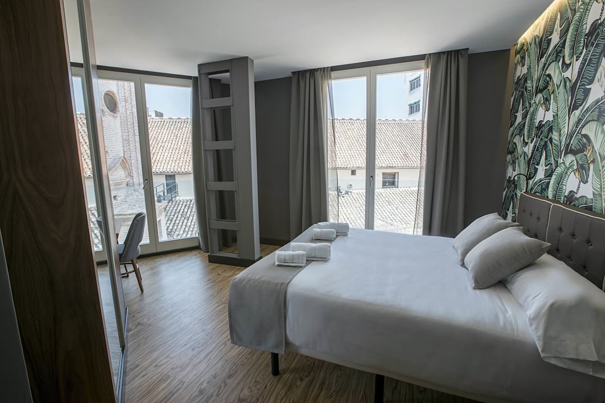 Habitación de hotel con una cama grande, una ventana con vistas a edificios, suelo de madera y una silla.