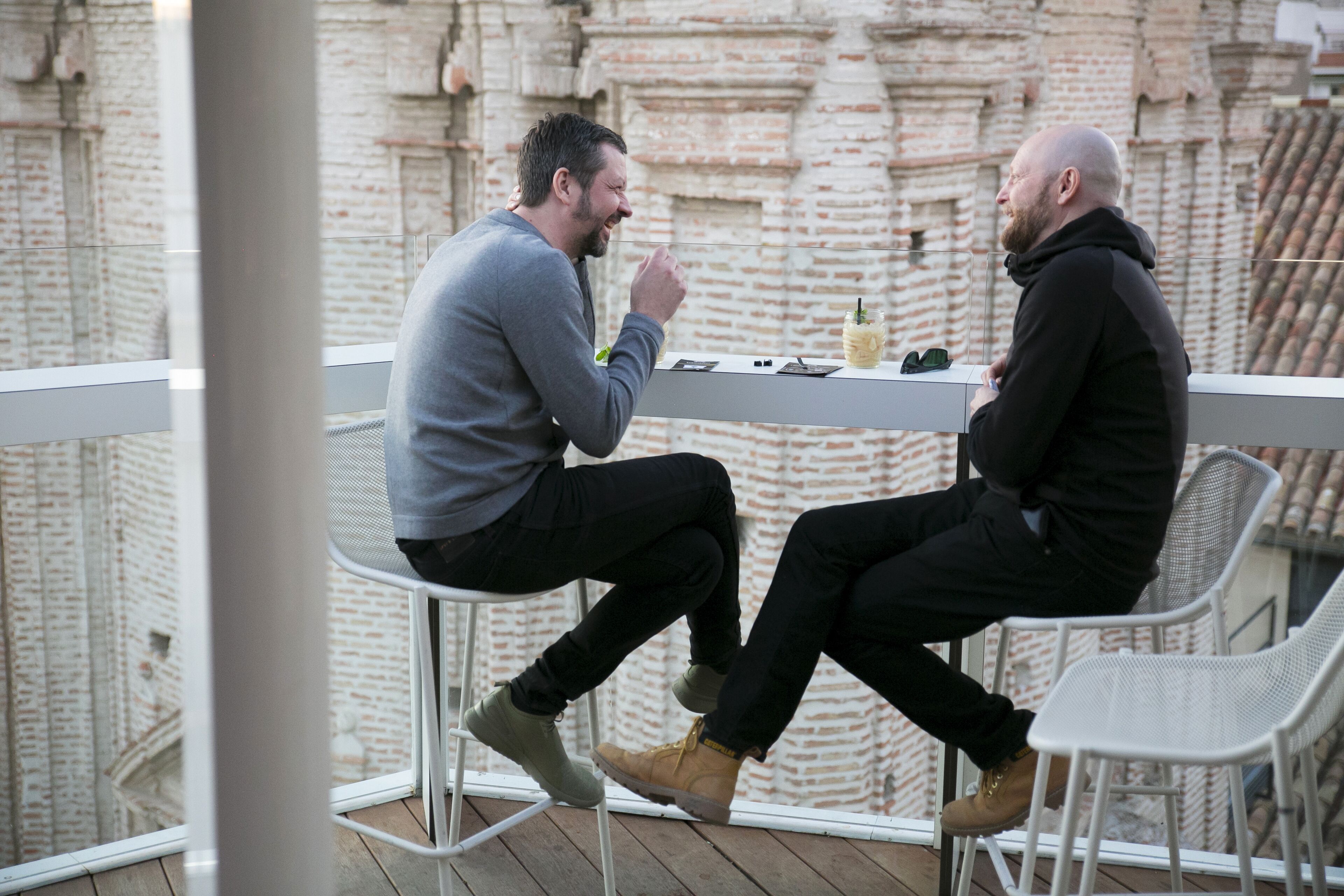 Dos hombres sentados en una mesa en un balcón con un muro de ladrillo al fondo.