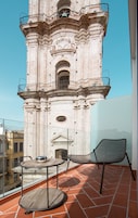 A balcony with a round wooden table and a metal chair, overlooking a historic brick tower with a balcony.