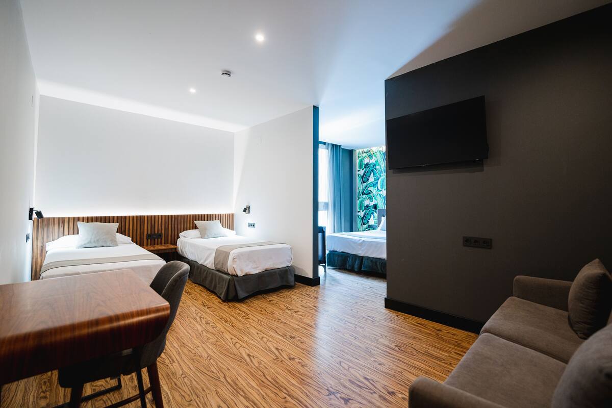 Una habitación de hotel moderna con dos camas, piso de madera, televisor de pantalla plana y un área de descanso con mesa y sofá.