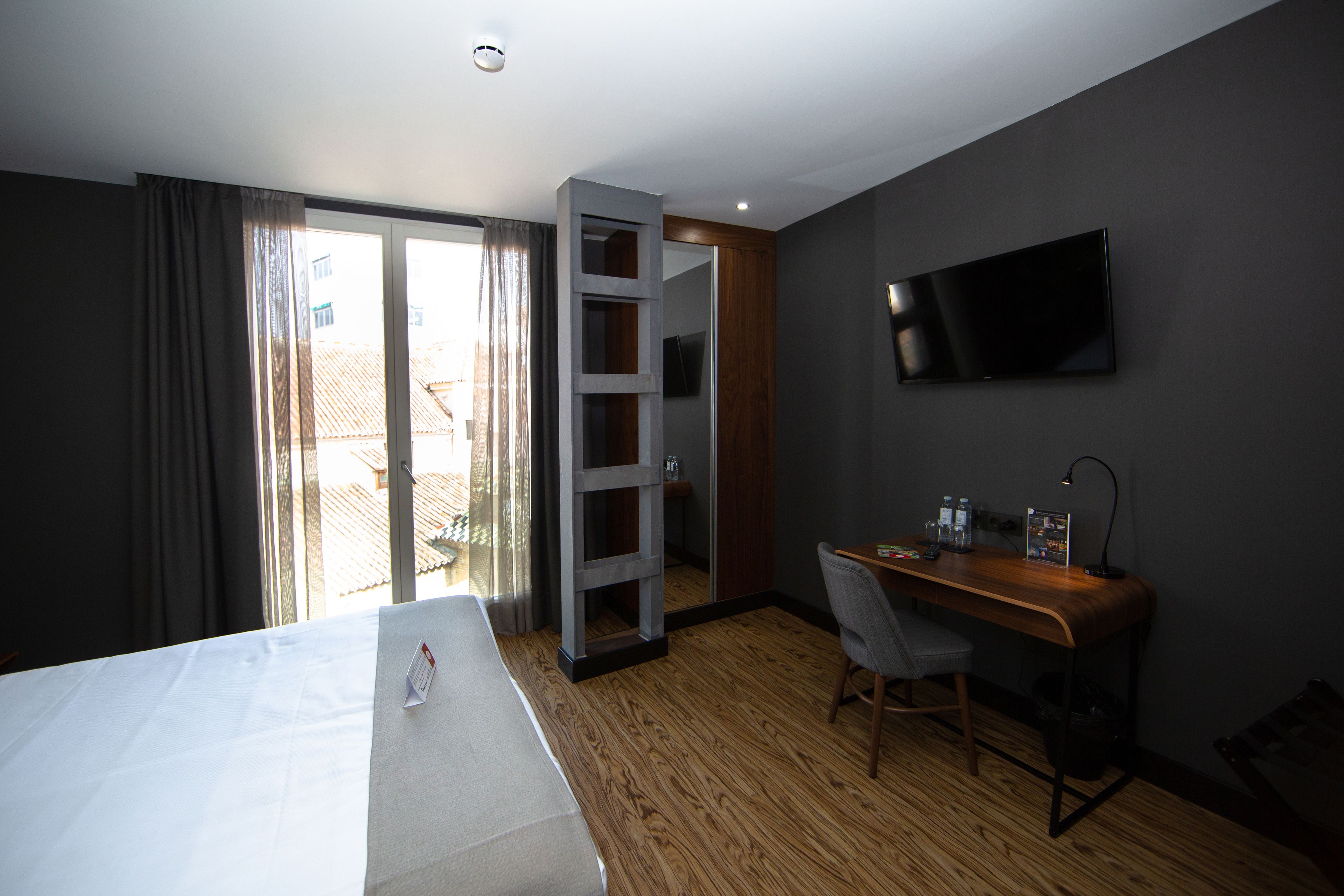 Habitación de hotel con cama, escritorio, silla, televisor y puerta corrediza a un balcón.