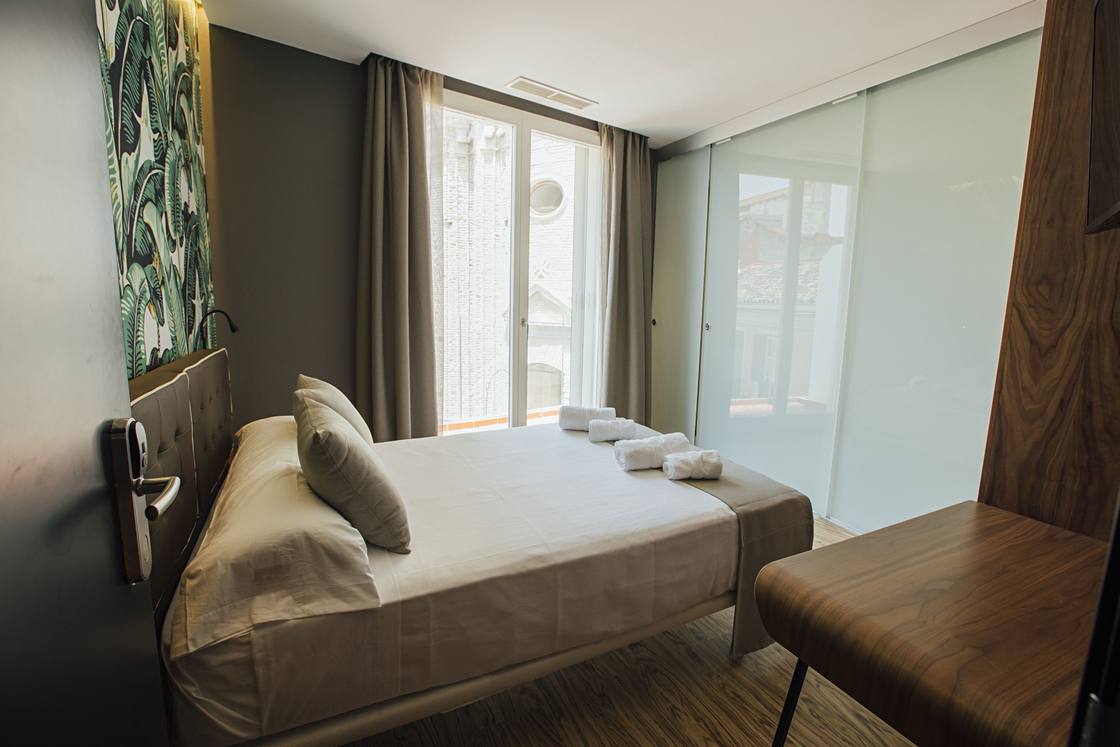 A hotel room with a bed, a wooden chair, a window with curtains, and a door with a handle.