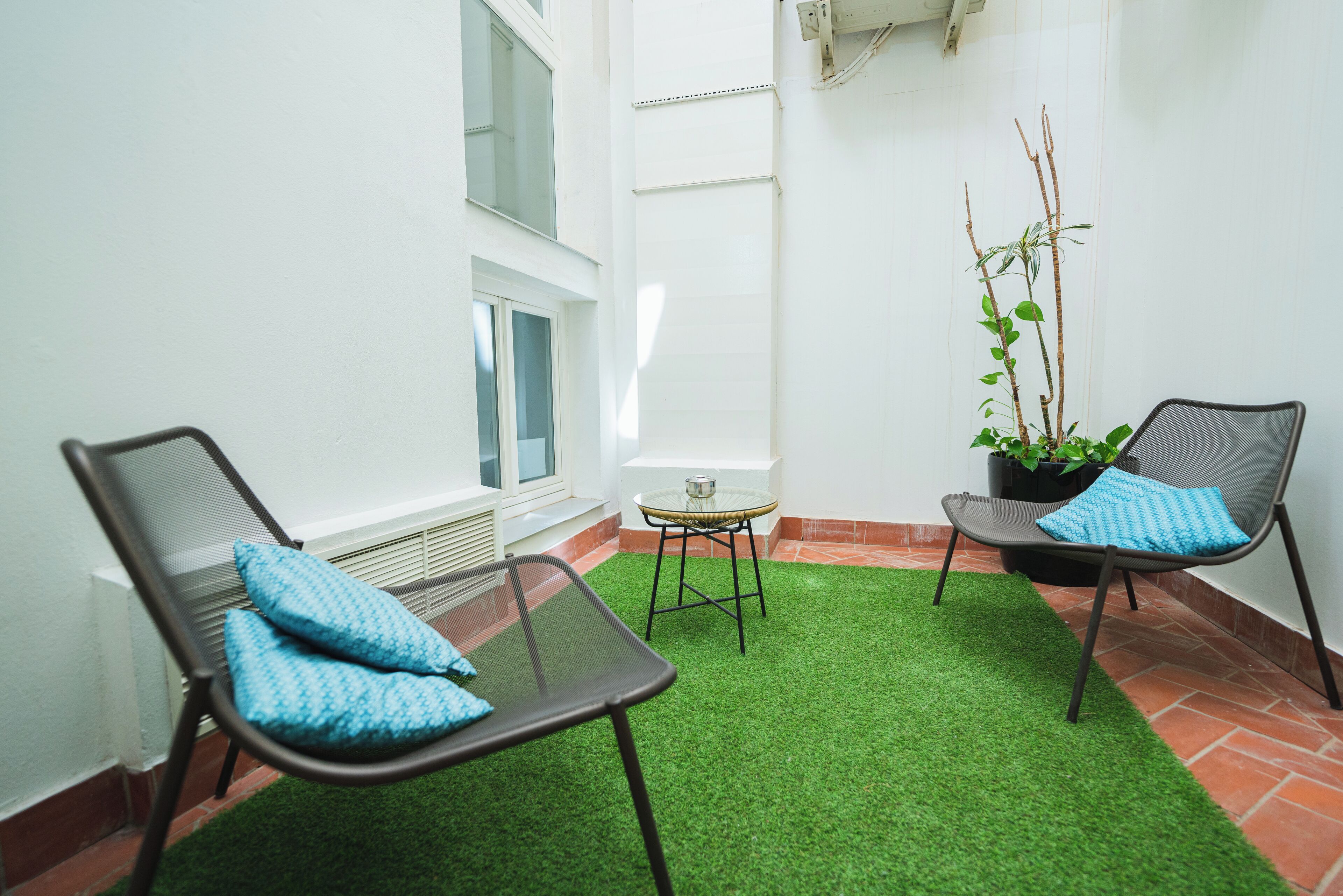 A small patio area with two chairs and a table, surrounded by white walls and a green artificial turf floor.