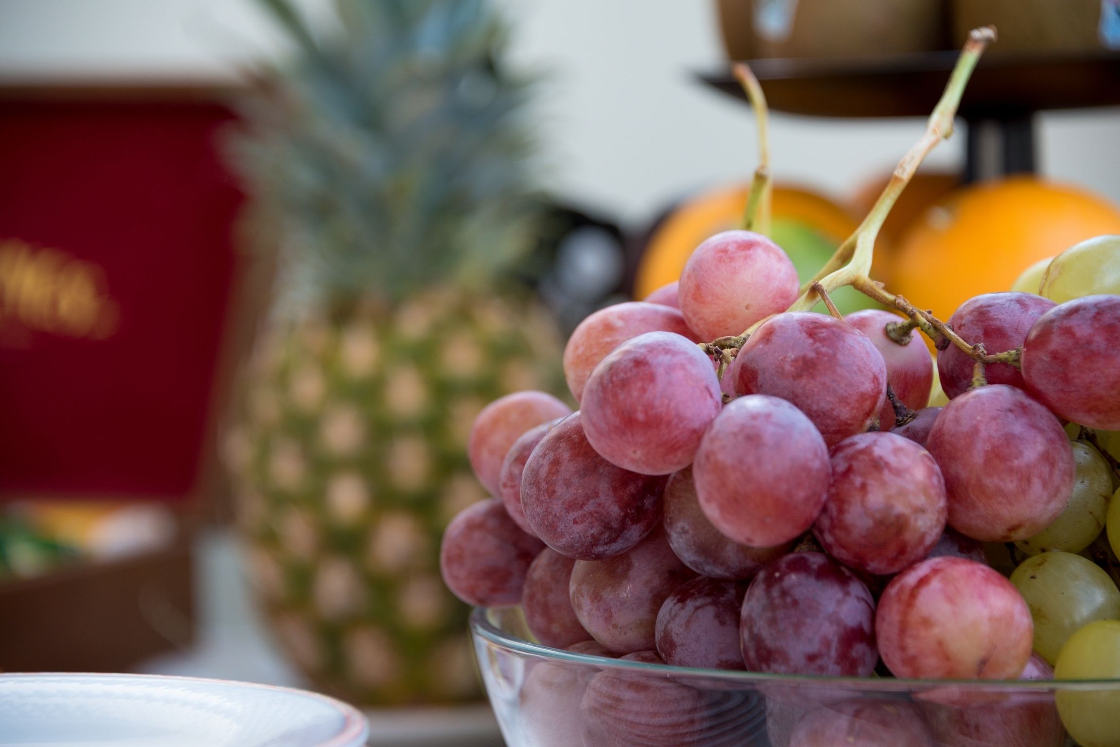 A bowl of red grapes with a pineapple and an orange in the background.