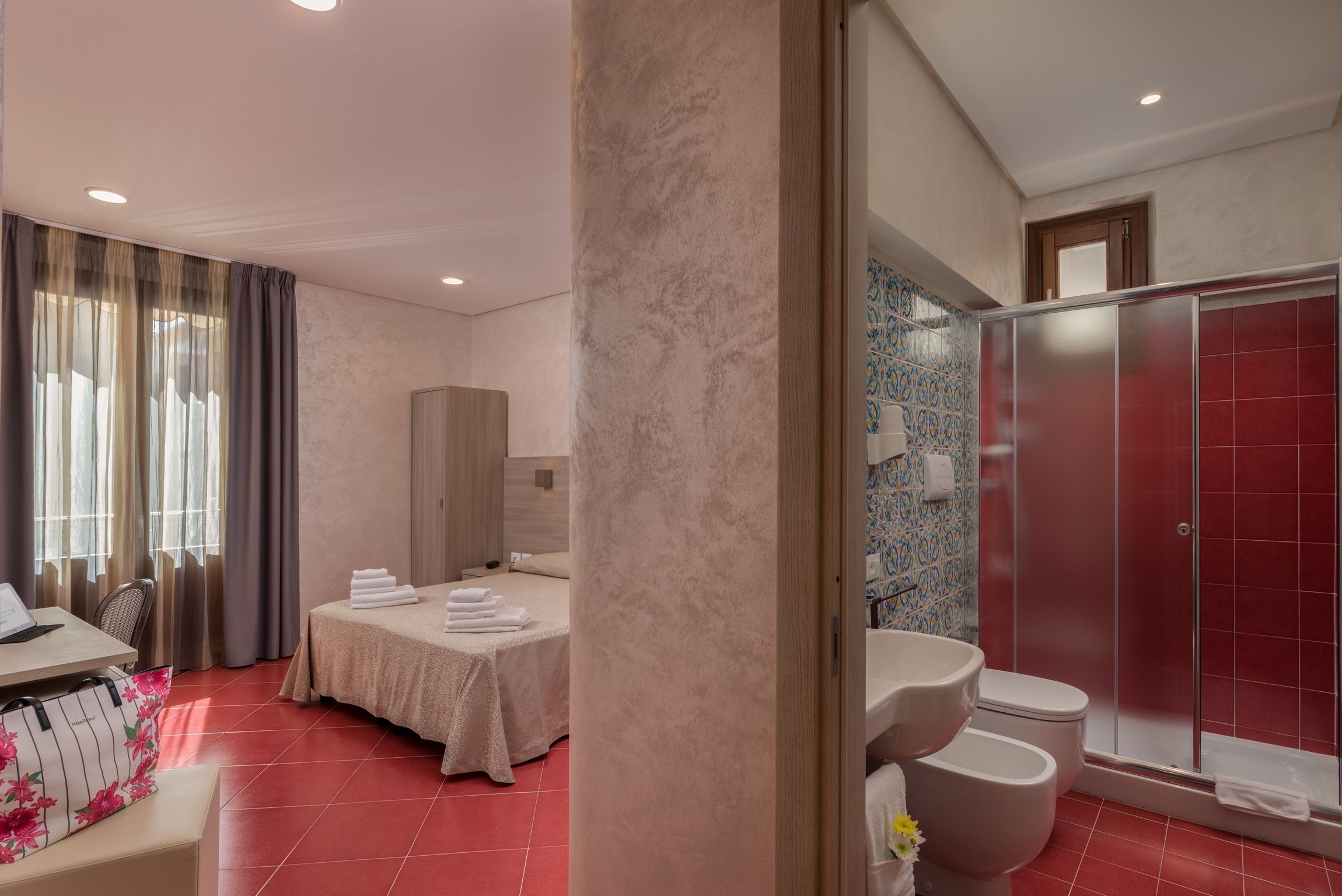 A hotel room with a bed, a desk with a laptop, a bathroom with a shower and toilet, and a window with curtains.