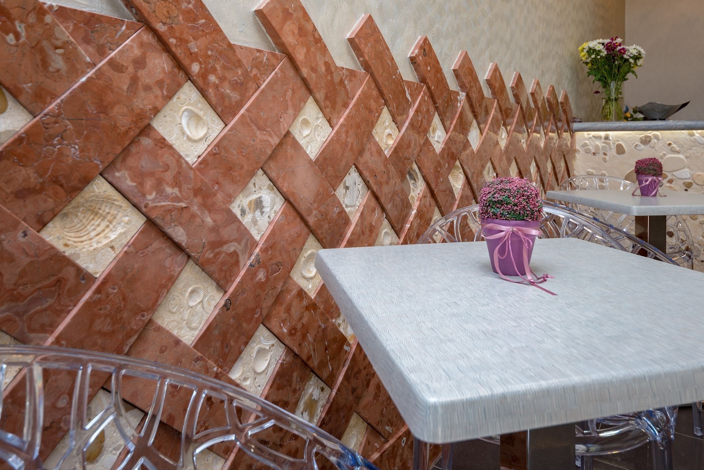 A restaurant interior with a patterned wall, clear glass chairs, and a table with a pink flower arrangement.