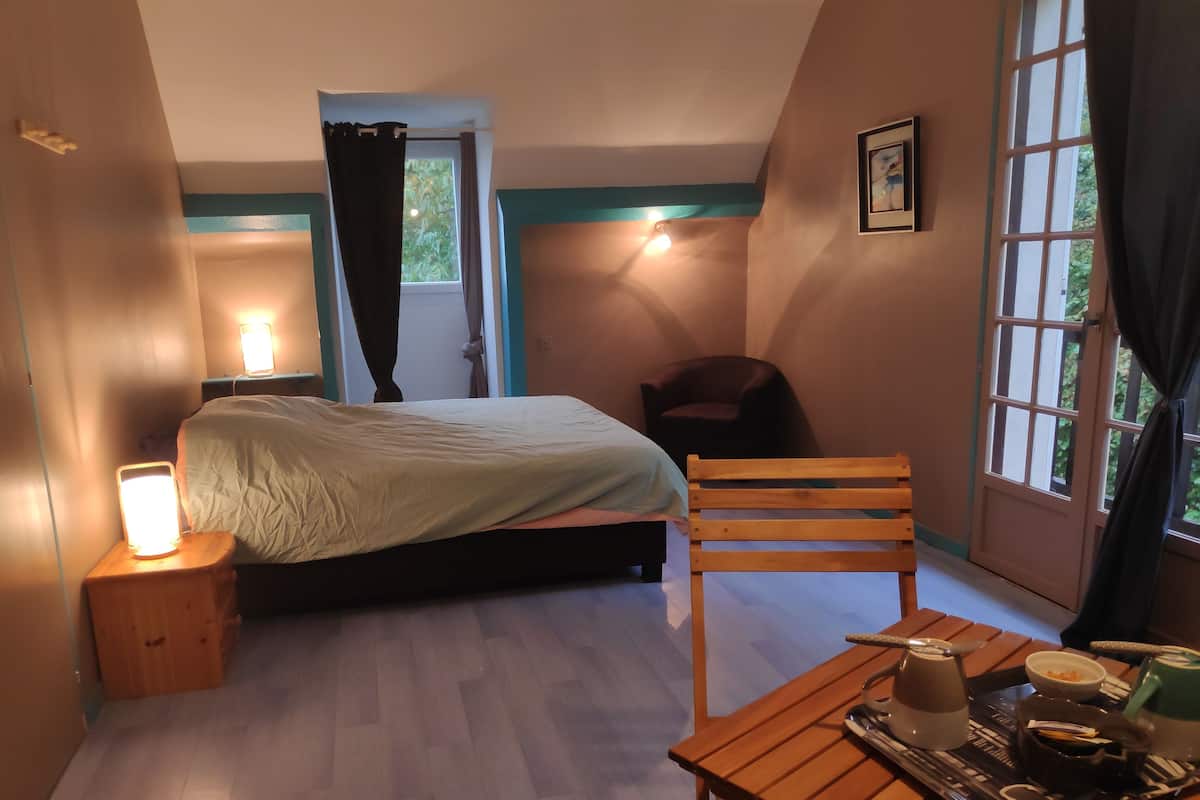 Une chambre à coucher avec un lit, une chaise, une table avec une bouilloire et une fenêtre avec des rideaux.