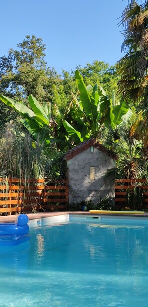 Une piscine avec une petite construction en arrière-plan qui ressemble à une maison, le tout entouré d’une végétation luxuriante et d’une clôture en bois.