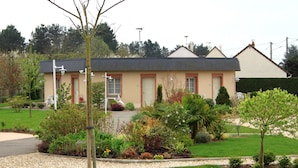 Een gebouw met twee verdiepingen, een beige gevel en rode raamkozijnen, omgeven door een verzorgde tuin met diverse planten en een grindpad.