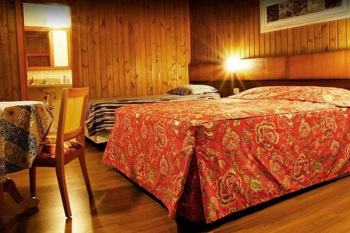 Une pièce aux murs en bois, un lit recouvert d’une couverture à motifs rouges, une chaise avec une nappe à motifs et une table de chevet avec une lampe.