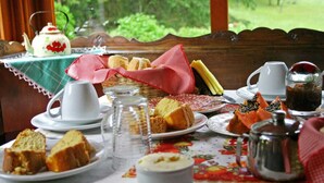 A table set with various food items and tea, including bread, pastries, and a teapot, with a view of greenery outside. - Chalets do Vale (Gramado)