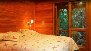 A wooden cabin bedroom with a bed, a nightstand, and a window with a view of the forest. - Chalets do Vale (Gramado)