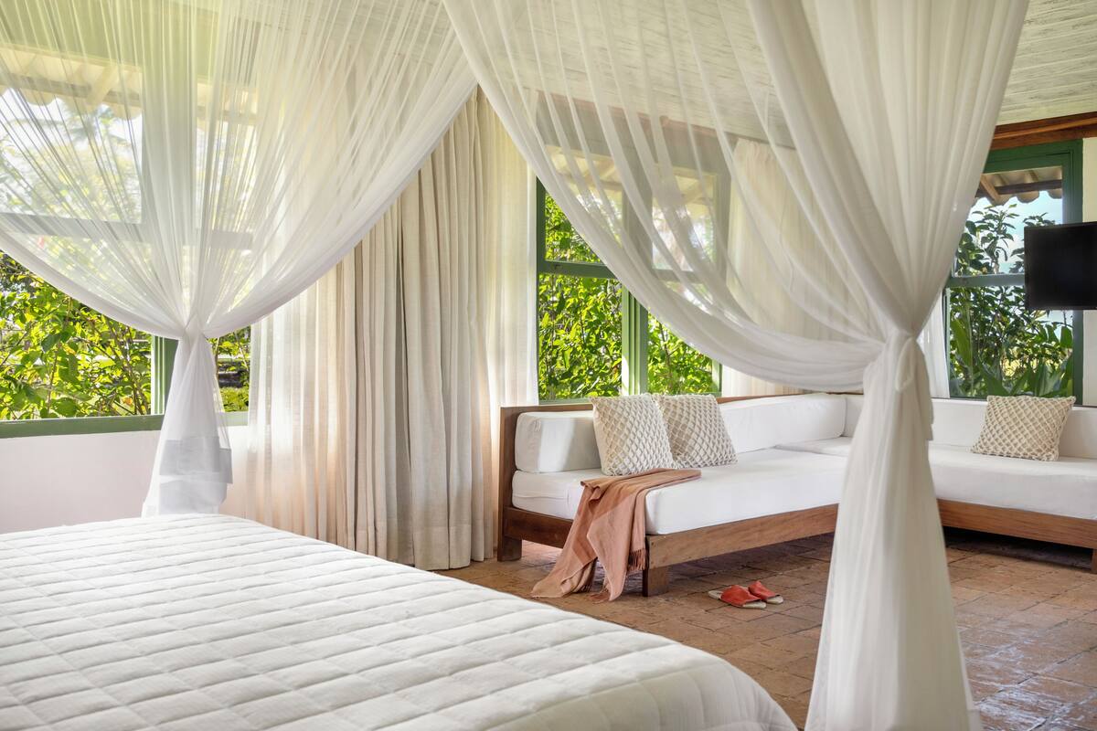A bedroom with a bed, a sofa, sheer curtains, and a view of greenery outside.
