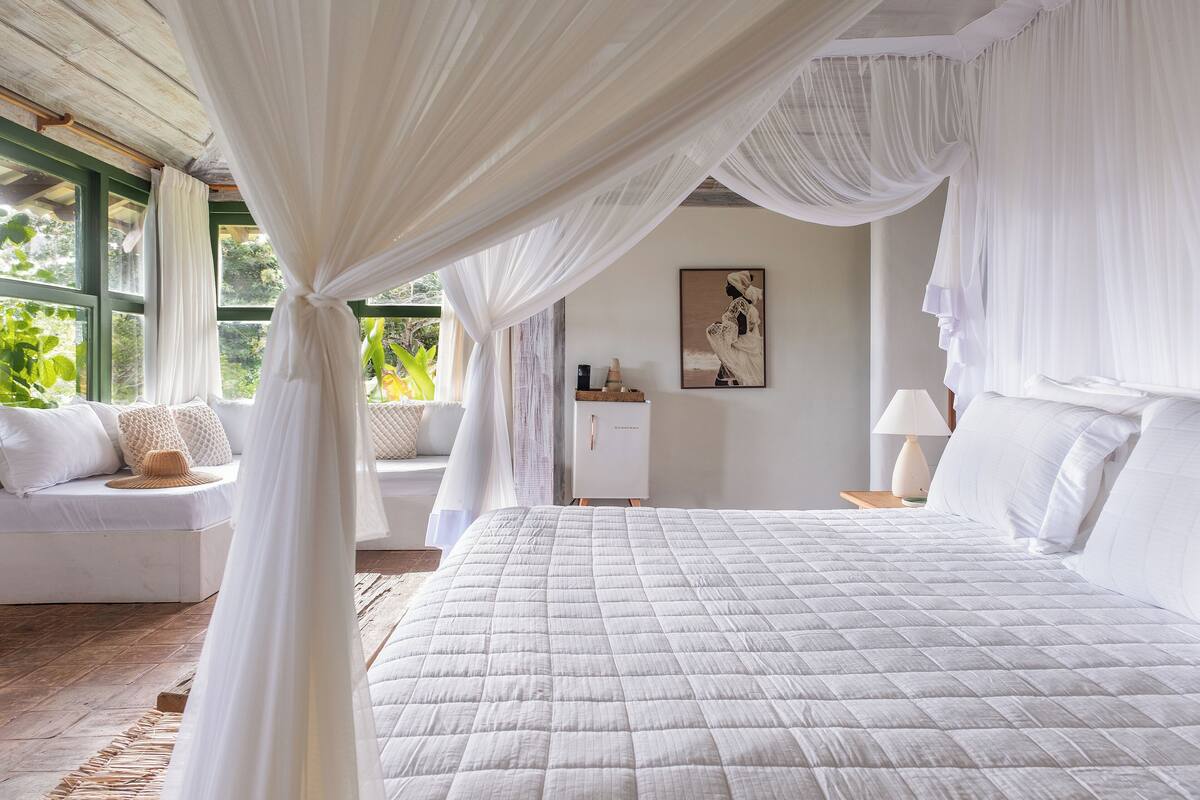 A bedroom with a large bed, white curtains, a sofa, and a nightstand with a lamp.