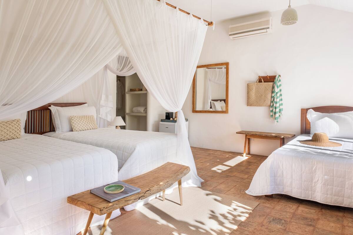 A bedroom with two beds, a wooden bench, a bedside table, a mirror, and a hanging basket.