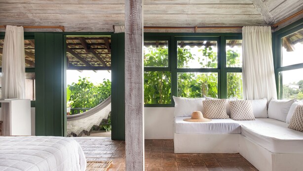 A room with a bed, a white sofa with cushions, a hammock, and large windows with green frames. - Pousada Outeiro (Porto Seguro)