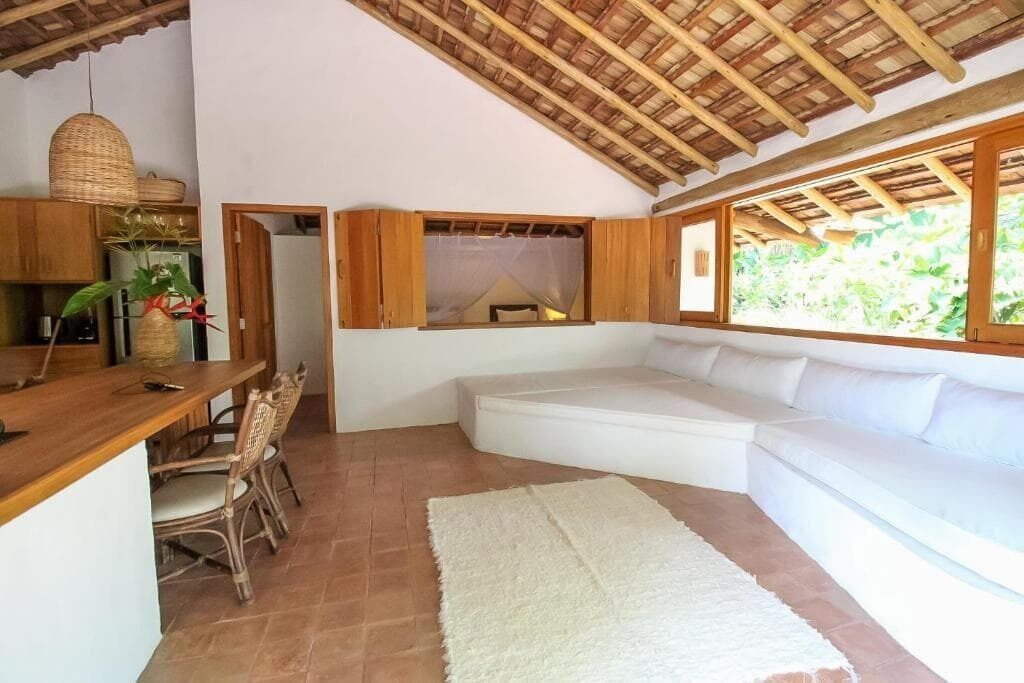 A room with a white sofa, a dining table with chairs, a kitchen area, and a large window with a view of greenery.