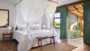 A bedroom with a four-poster bed, a bedside table with a lamp, and a view of the outdoors through a window. - Pousada Outeiro (Porto Seguro)