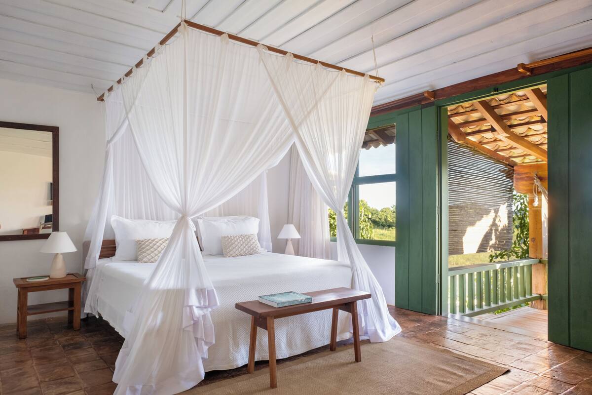 A bedroom with a four-poster bed, a bedside table with a lamp, and a view of the outdoors through a window.