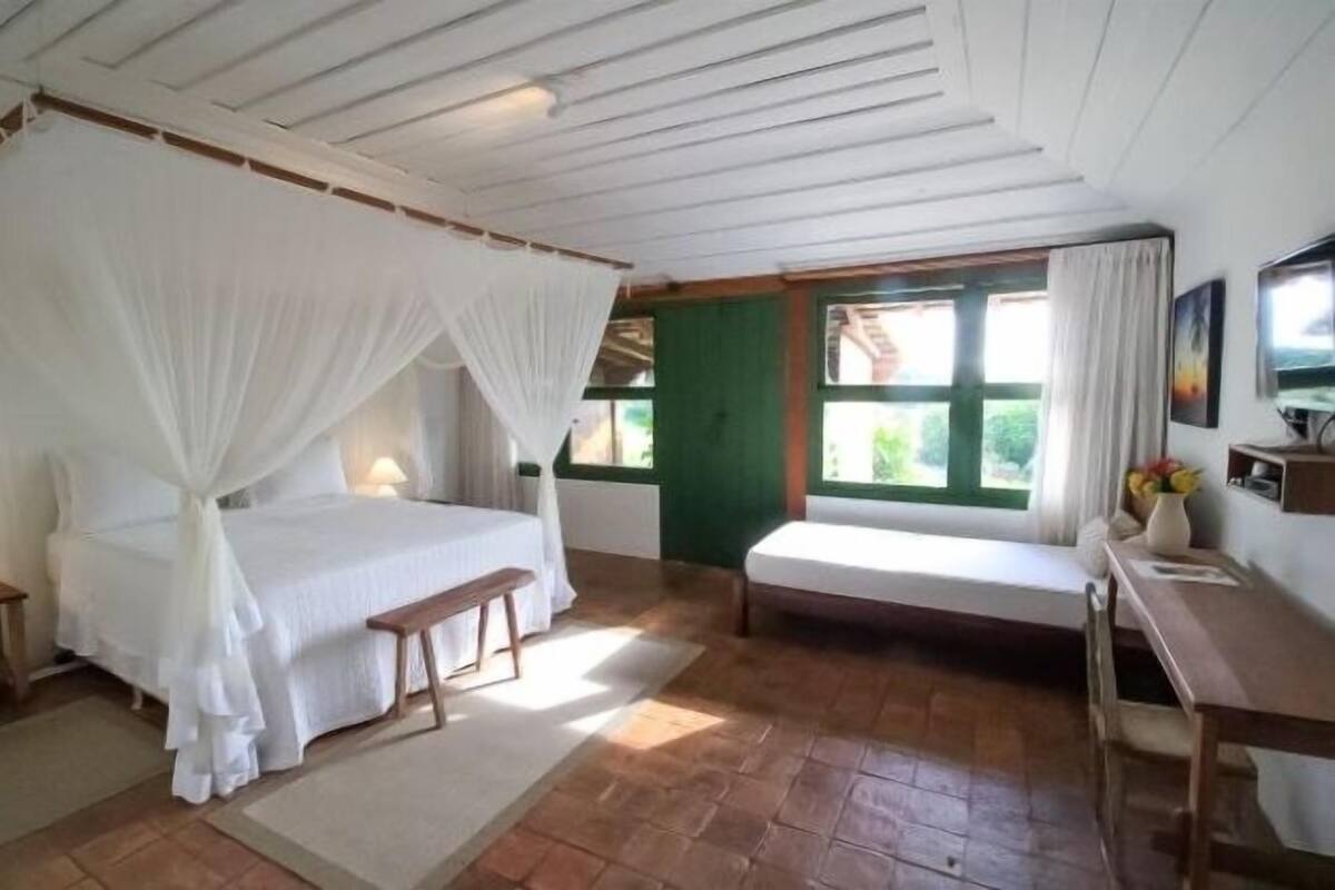 A bedroom with a bed under a canopy, a bench, a desk, and a window with curtains.