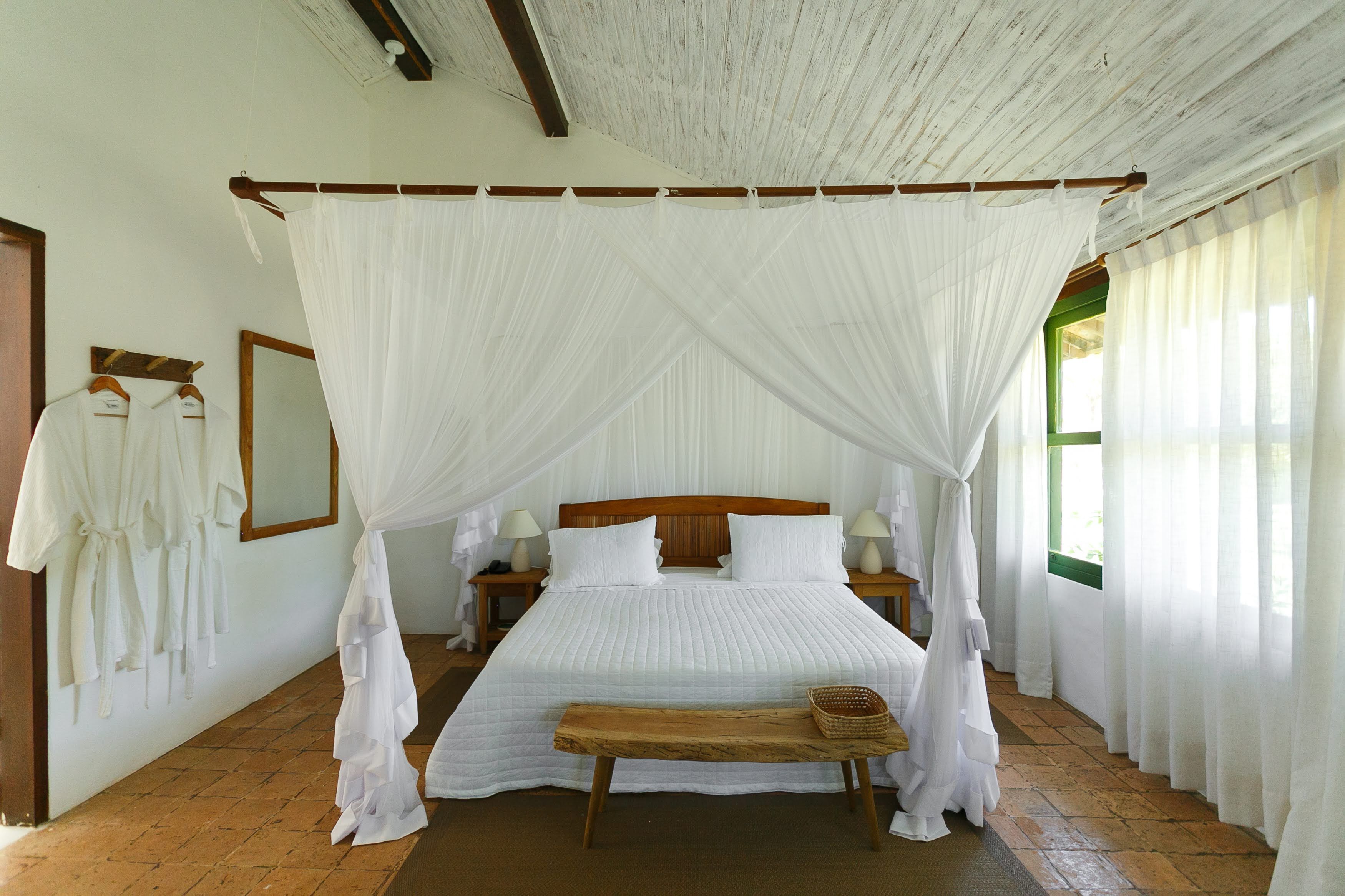 Habitación con cama bajo un dosel, cabecero de madera, mesitas de noche con lámparas, un banco y una ventana con cortinas.