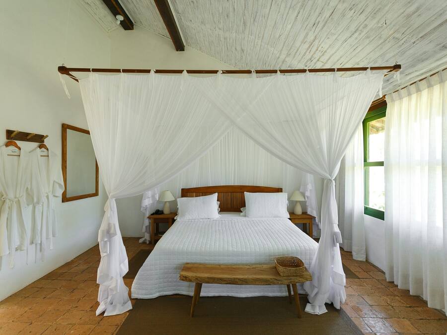 Habitación con cama bajo un dosel, cabecero de madera, mesitas de noche con lámparas, un banco y una ventana con cortinas.