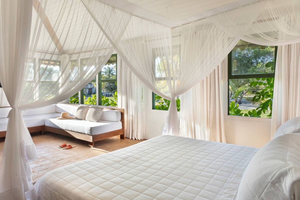 A bedroom with a large bed, a sofa, and a window with a view of greenery.