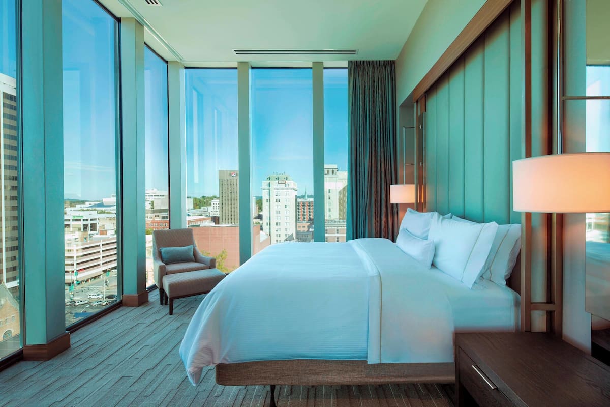 A hotel room with a large bed, a chair, a nightstand, and a view of the city through large windows.