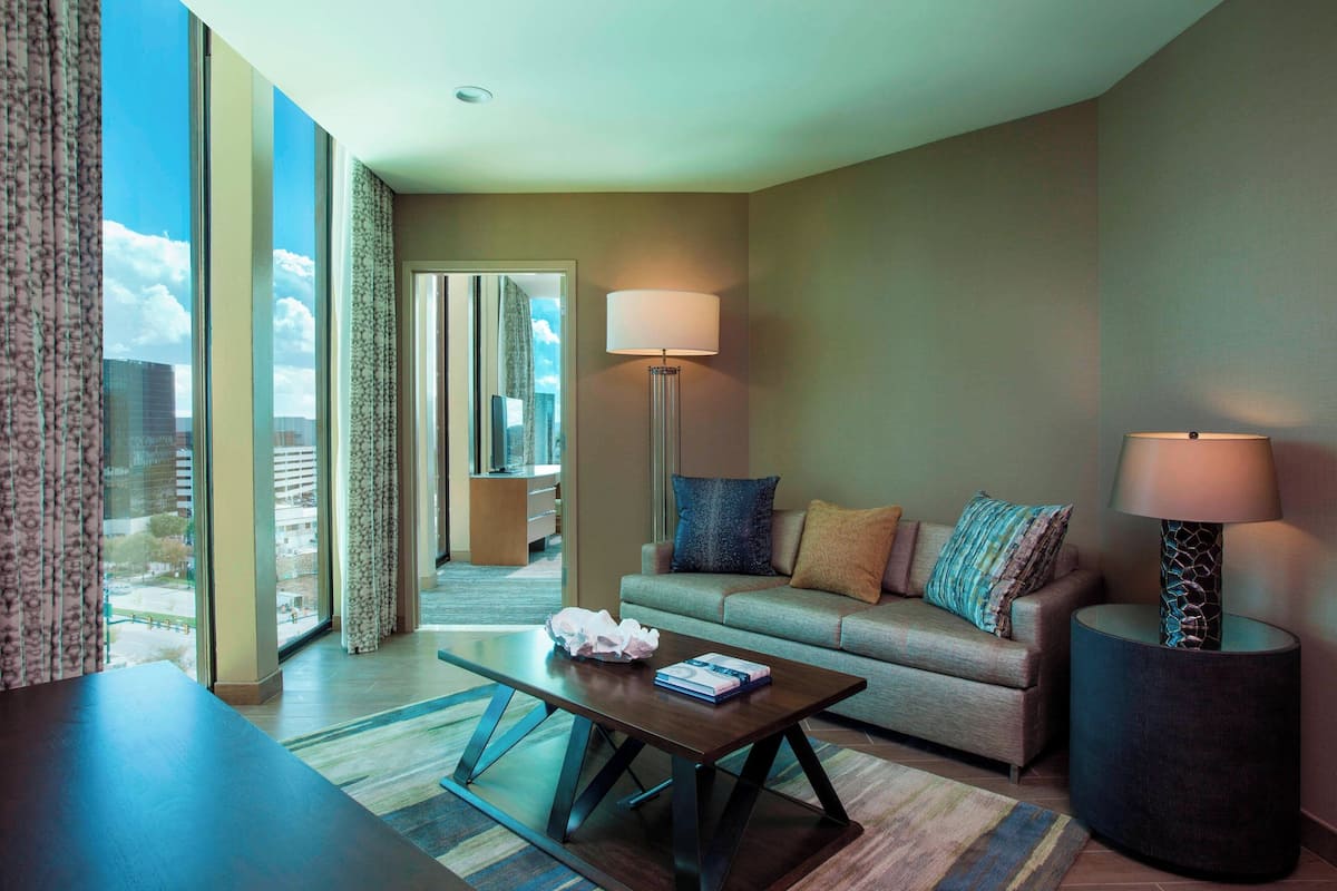 A modern hotel room with a large window, a sofa, a coffee table, and two floor lamps.