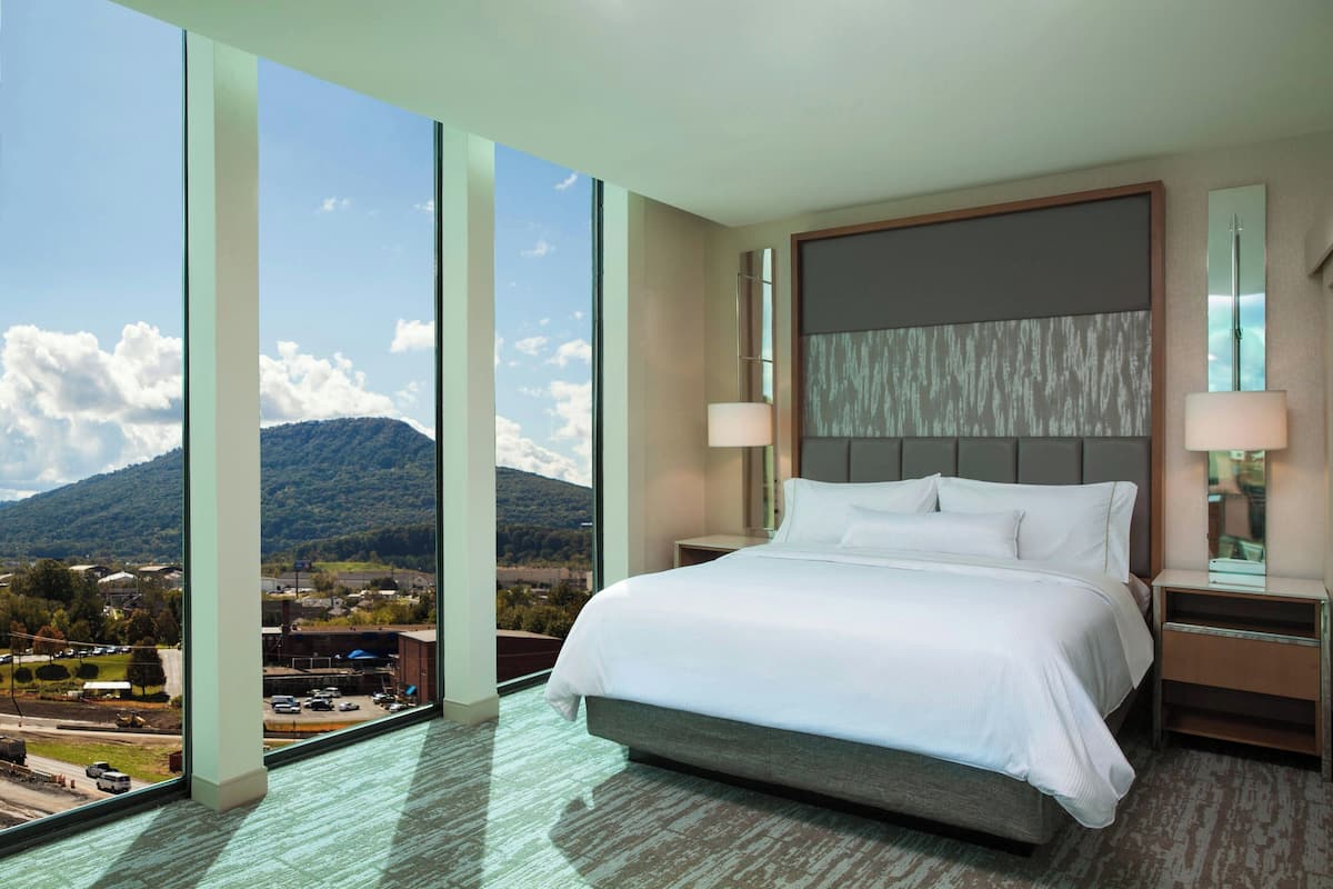 A hotel room with a large bed, two bedside lamps, a nightstand, and a view of a mountainous landscape through large windows.