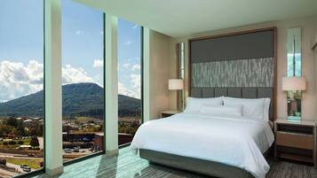Habitación de hotel con una cama grande, dos lámparas de noche, una mesita de noche y vistas a un paisaje montañoso a través de amplios ventanales.