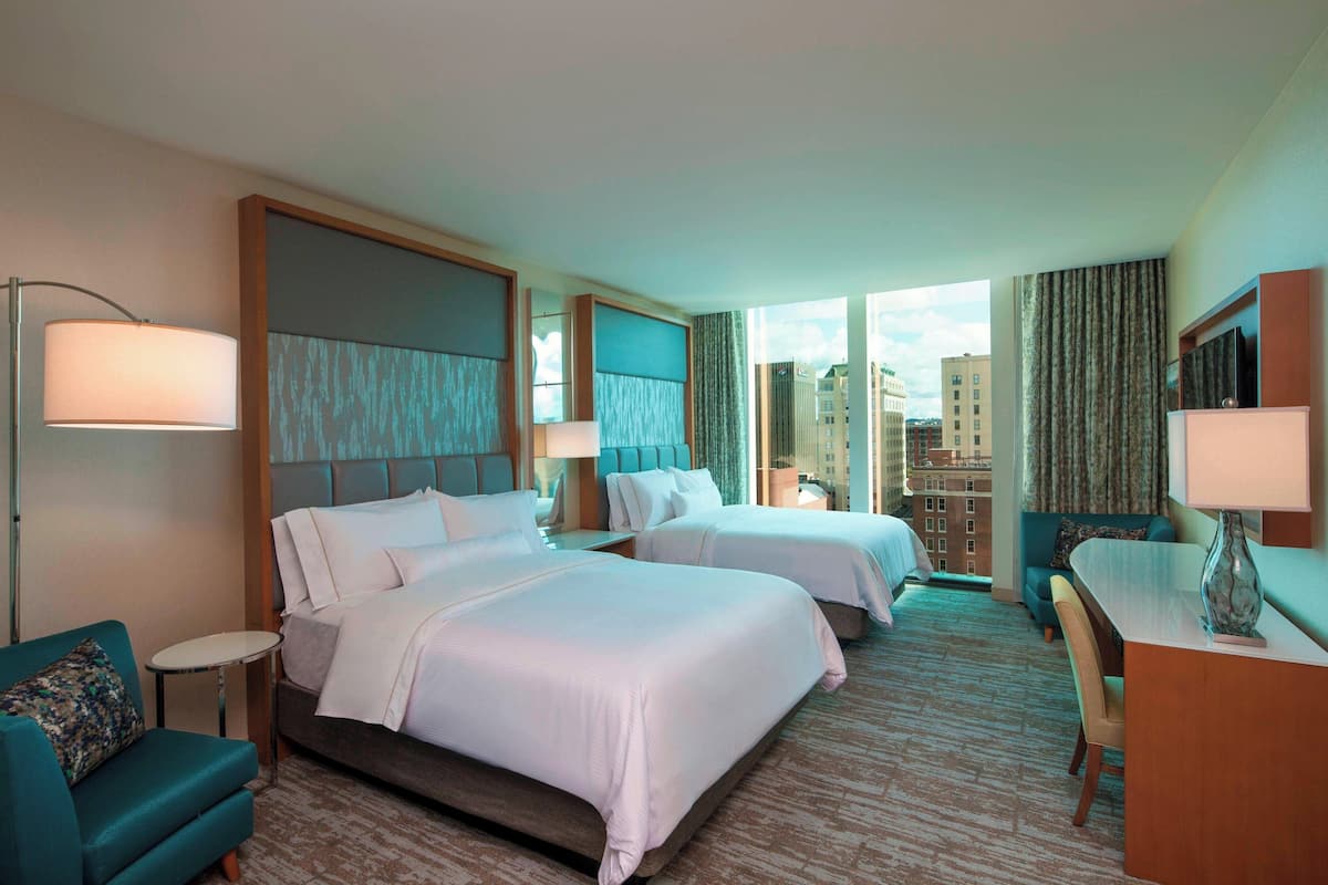 A hotel room with two beds, a desk, a chair, a small table, and a view of the city.