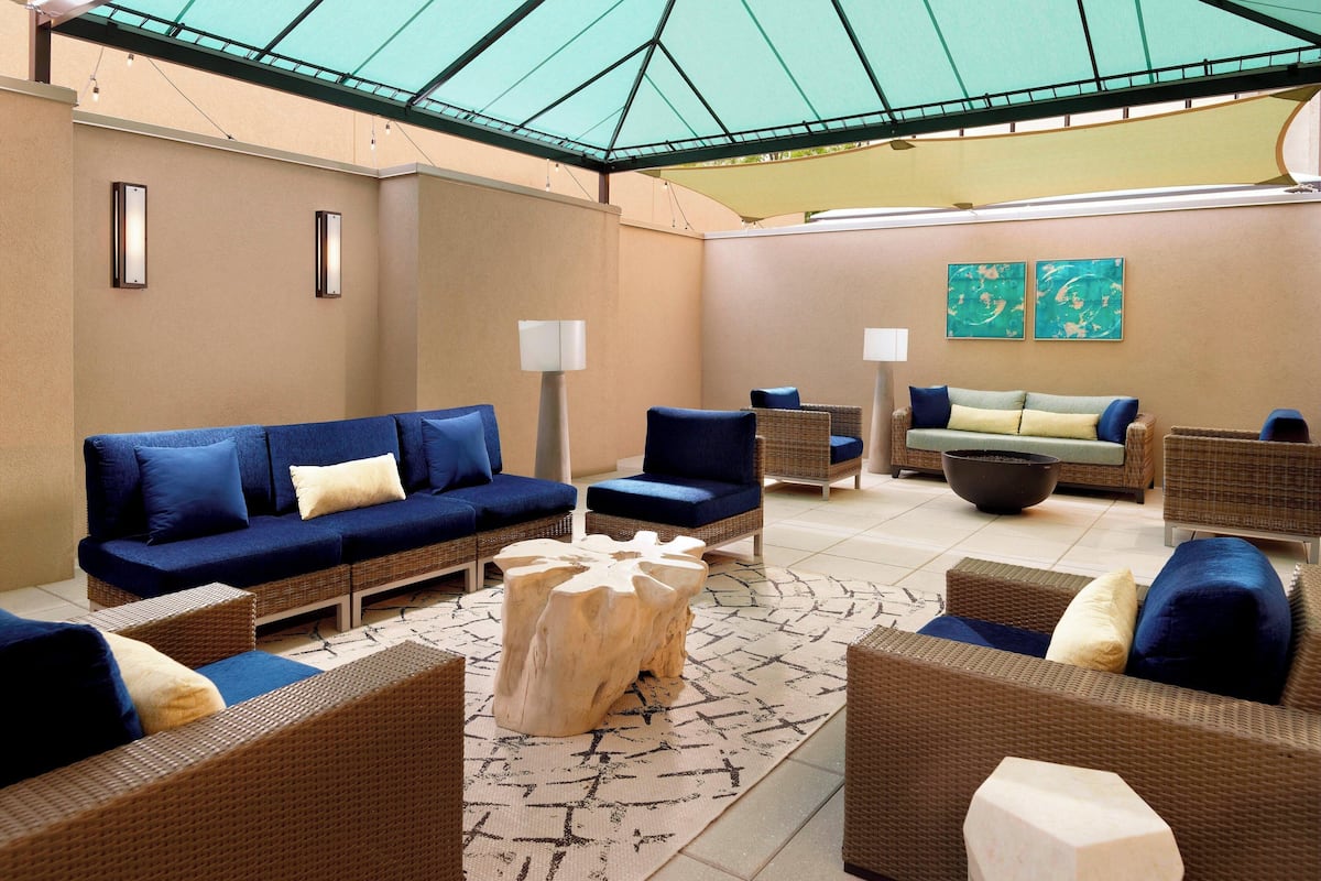 A sunlit outdoor lounge area with wicker furniture, a central wooden coffee table, and two wall-mounted lamps.