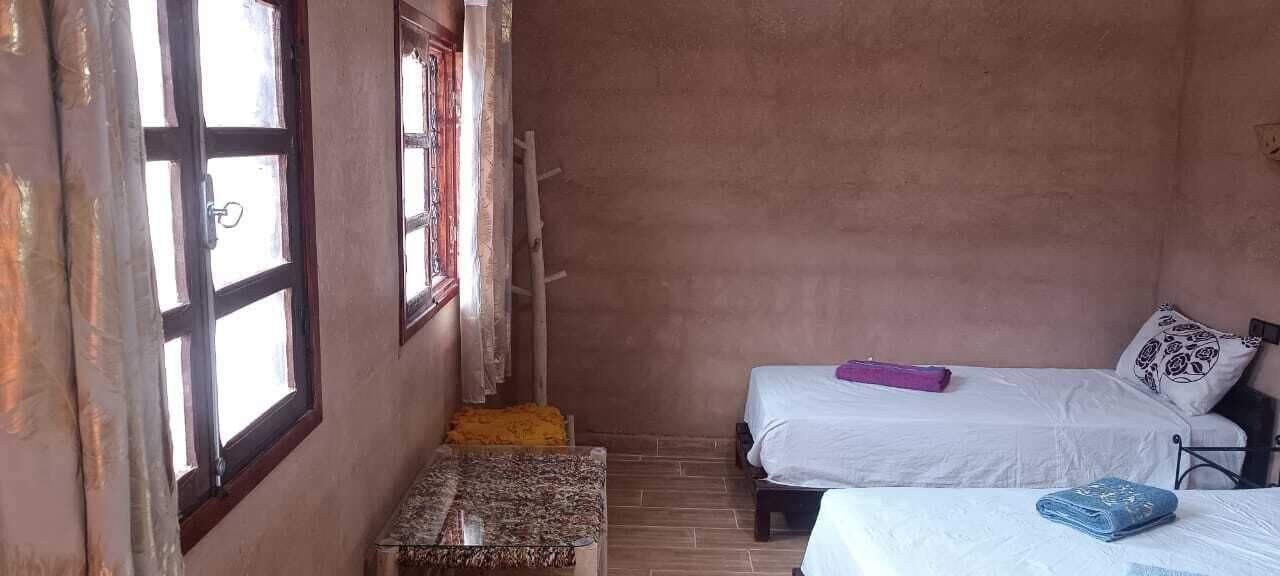 Una habitación pequeña y rústica con dos camas, un banco de madera y una ventana con cortinas.