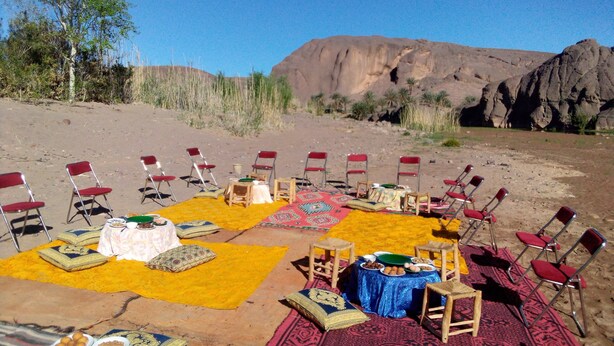 Un espace de restauration en plein air, avec des chaises et des tables disposées sur des tapis à motifs, le tout intégré dans un paysage désertique parsemé de formations rocheuses.