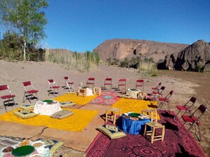 Een buiten eethoek met stoelen en tafels, gerangschikt op kleurrijke vloerkleden, met op de achtergrond een woestijnlandschap en rotsformaties.
