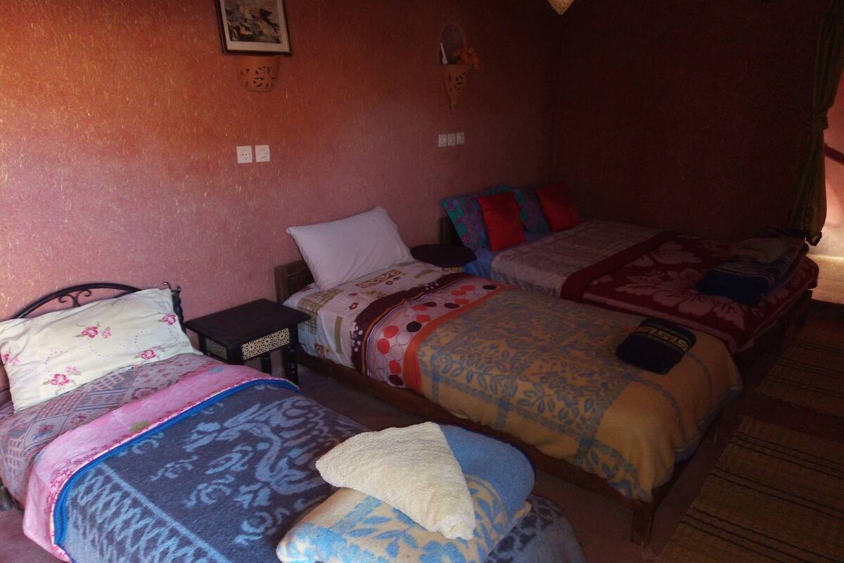 Ein Zimmer mit drei Betten, jedes mit unterschiedlicher Bettwäsche. Es gibt Steckdosen und eine kleine, an der Wand befestigte Ablage mit einem dekorativen Gegenstand.