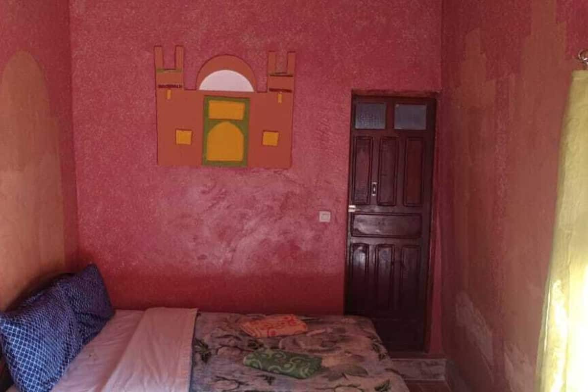 Ein kleiner, schmaller Raum mit einem Einzelbett, einer Holztür und einem an der Wand befestigten Regal.